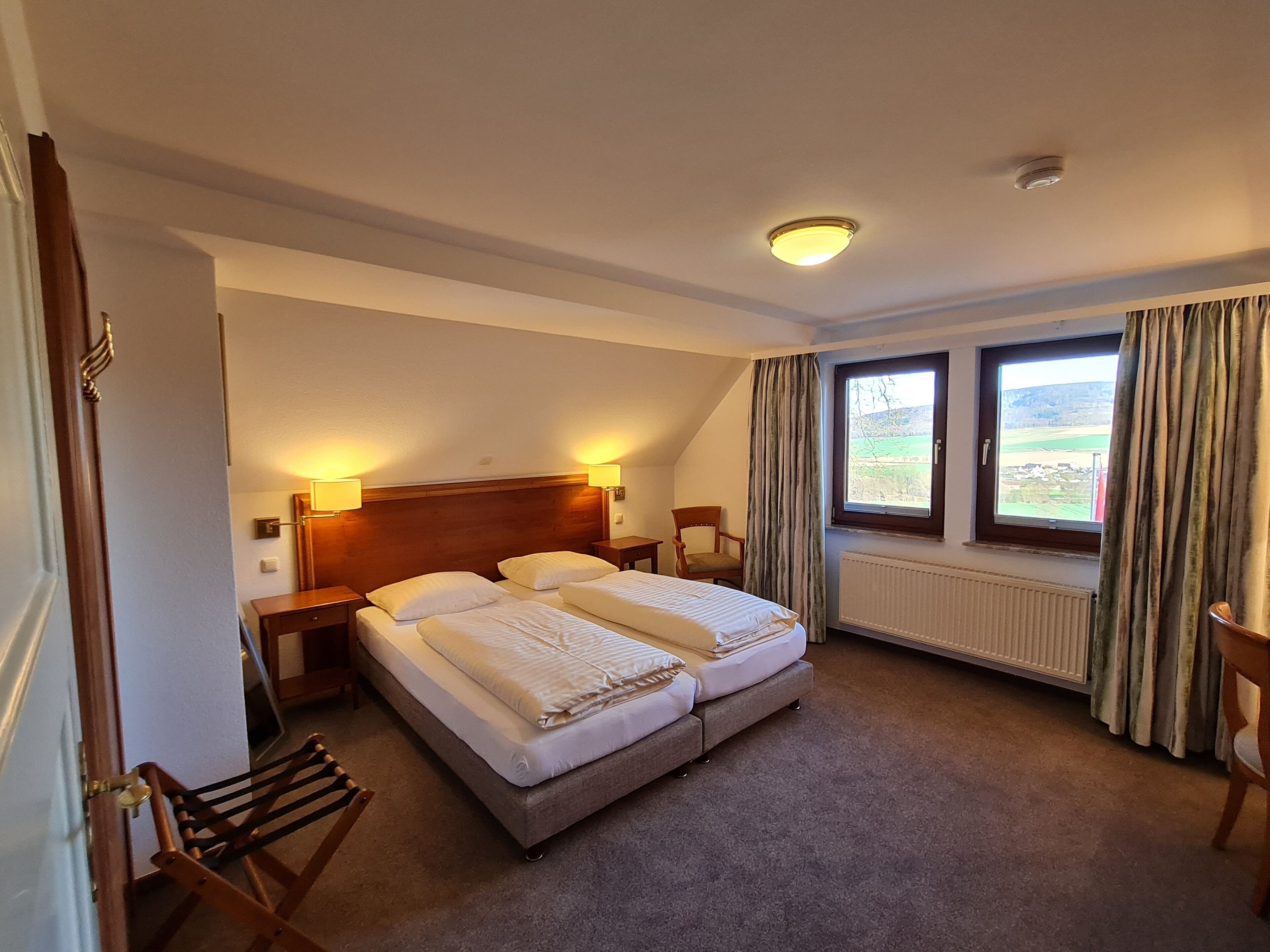 A bedroom with two beds, a wooden headboard, bedside lamps, a radiator, and a window with curtains.