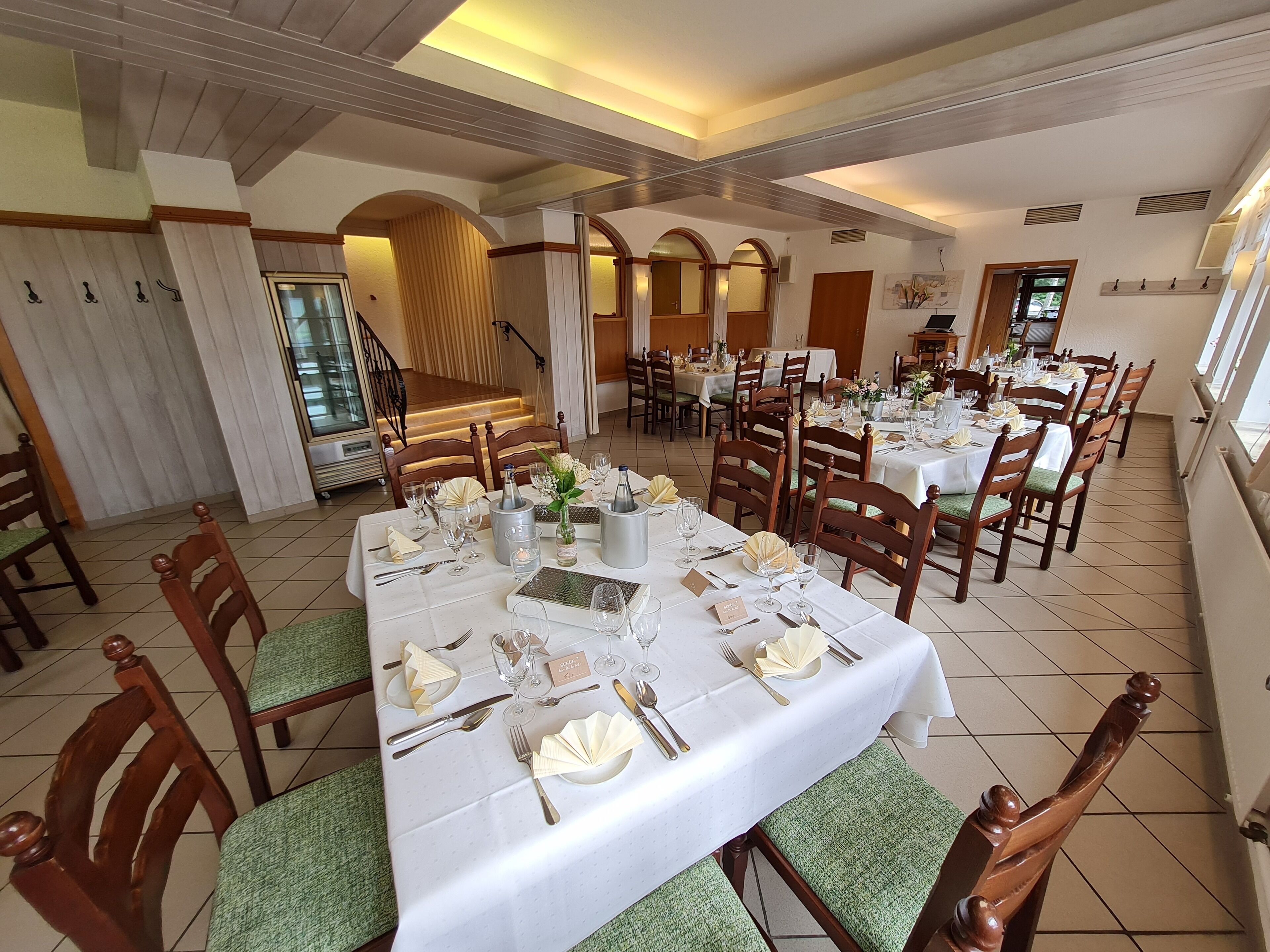 Un comedor amplio con varias mesas dispuestas para una comida, sillas de madera y una escalera que conduce a un nivel superior.
