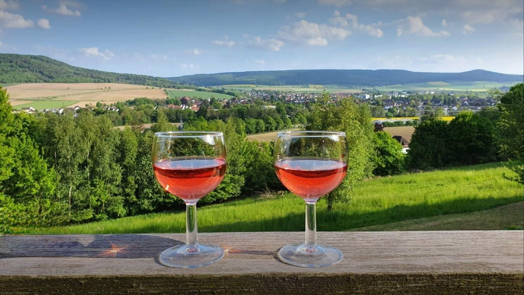 Dos copas de vino sobre una superficie de madera, con una vista panorámica de un pueblo y colinas al fondo.