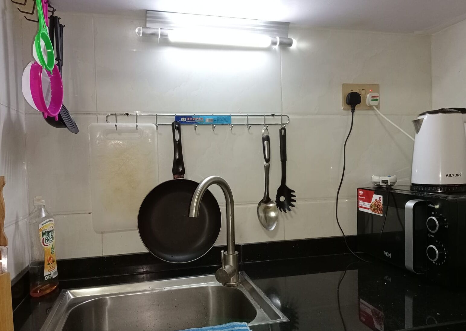 Een keuken met een roestvrijstalen spoelbak, een zwart aanrechtblad, een witte muur met een hangrek en een koffiezetapparaat.