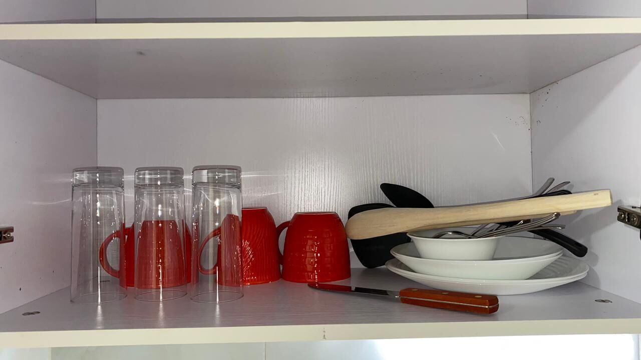 Une étagère de cuisine avec des tasses en verre transparent, des tasses rouges, un bol blanc, un couteau et une spatule en bois.