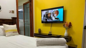 A hotel room with a bed, a TV mounted on the wall, a bedside table with a vase of flowers, and a wooden headboard. - Lux Suites Kileleshwa Studio Apartments (Nairobi)