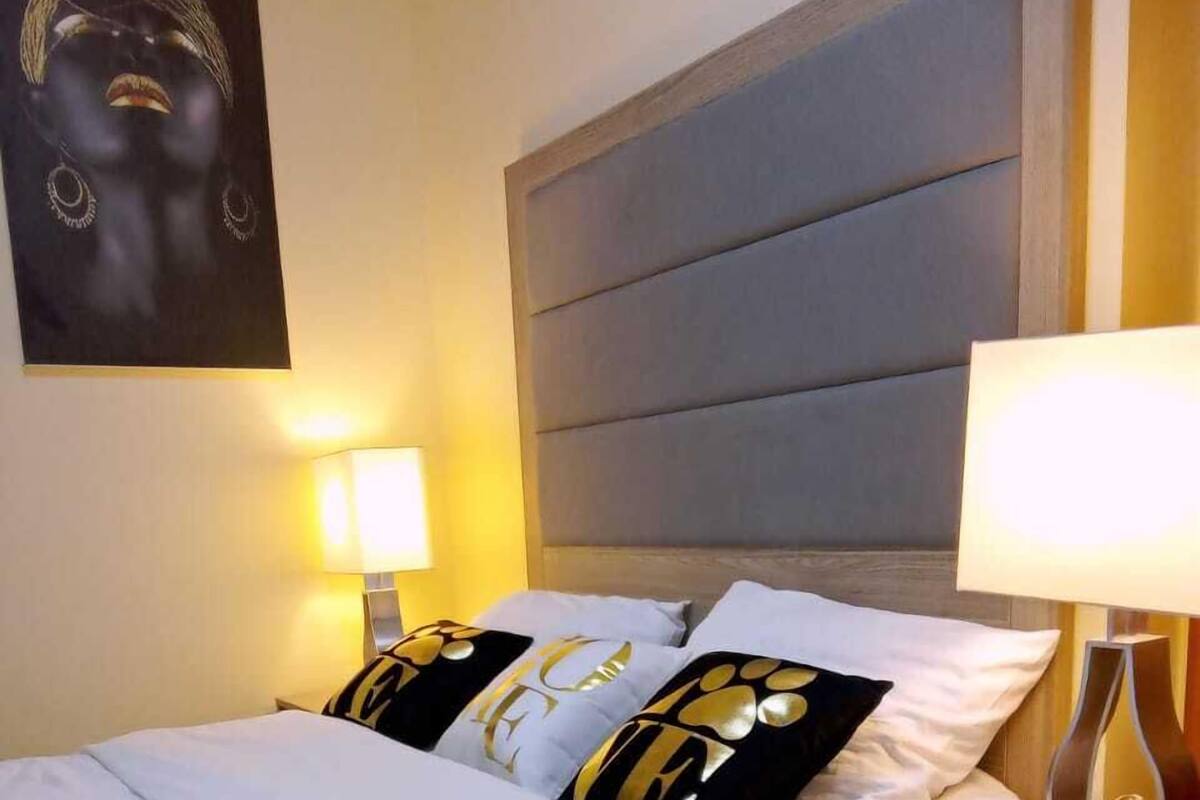 Un lit bien fait, agrémenté d’oreillers aux motifs blancs et dorés, d’une tête de lit rembourrée de couleur grise et de lampes de chevet.