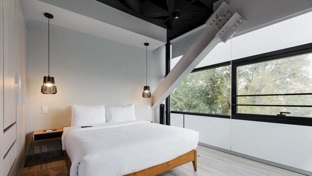 Une chambre moderne avec un grand lit, deux lampes suspendues, une table de chevet et une vue sur les arbres à travers une grande fenêtre.