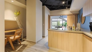Un intérieur moderne avec un bureau et une chaise en bois, un coin cuisine doté d’un comptoir, et une vue sur l’extérieur.