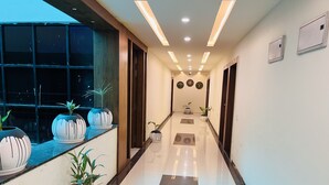 Un pasillo moderno de hotel con suelo reflectante, plantas en macetas, iluminación empotrada y vistas al cielo a través de grandes ventanales.