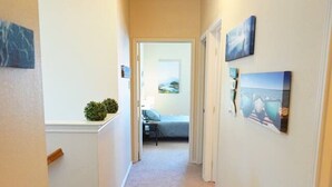 A hallway with beige walls, a carpeted floor, and a door leading to a bedroom. There are wall-mounted pictures and a small plant on the left side. - Cozy Home in Frisco/Little Elm (Little Elm)