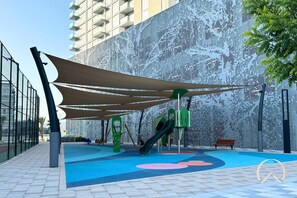 An outdoor playground with slides and a climbing structure, surrounded by a patterned fence and a large building in the background. - Vogue | District One Residences close to Downtown Dubai Mall | Burj Khalifa (Dubai)