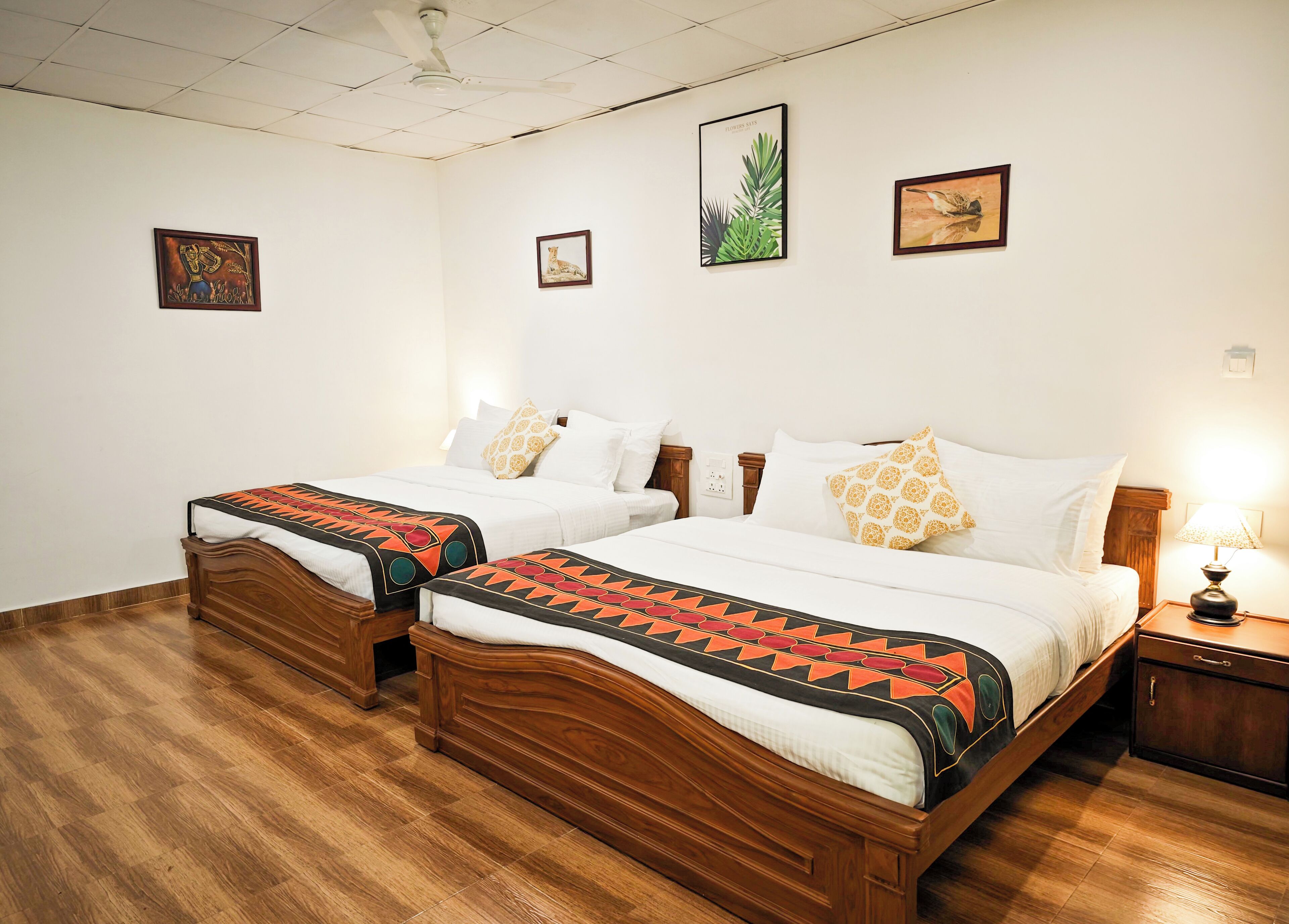 A hotel room with two wooden beds, white bedding, and a wooden floor. There are wall-mounted artworks and a nightstand with a lamp.