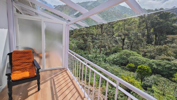 A balcony with a chair, white railings, and a view of a lush green landscape with trees and mountains. - Ibex Resorts, Valparai (Mistly) (Valparai)