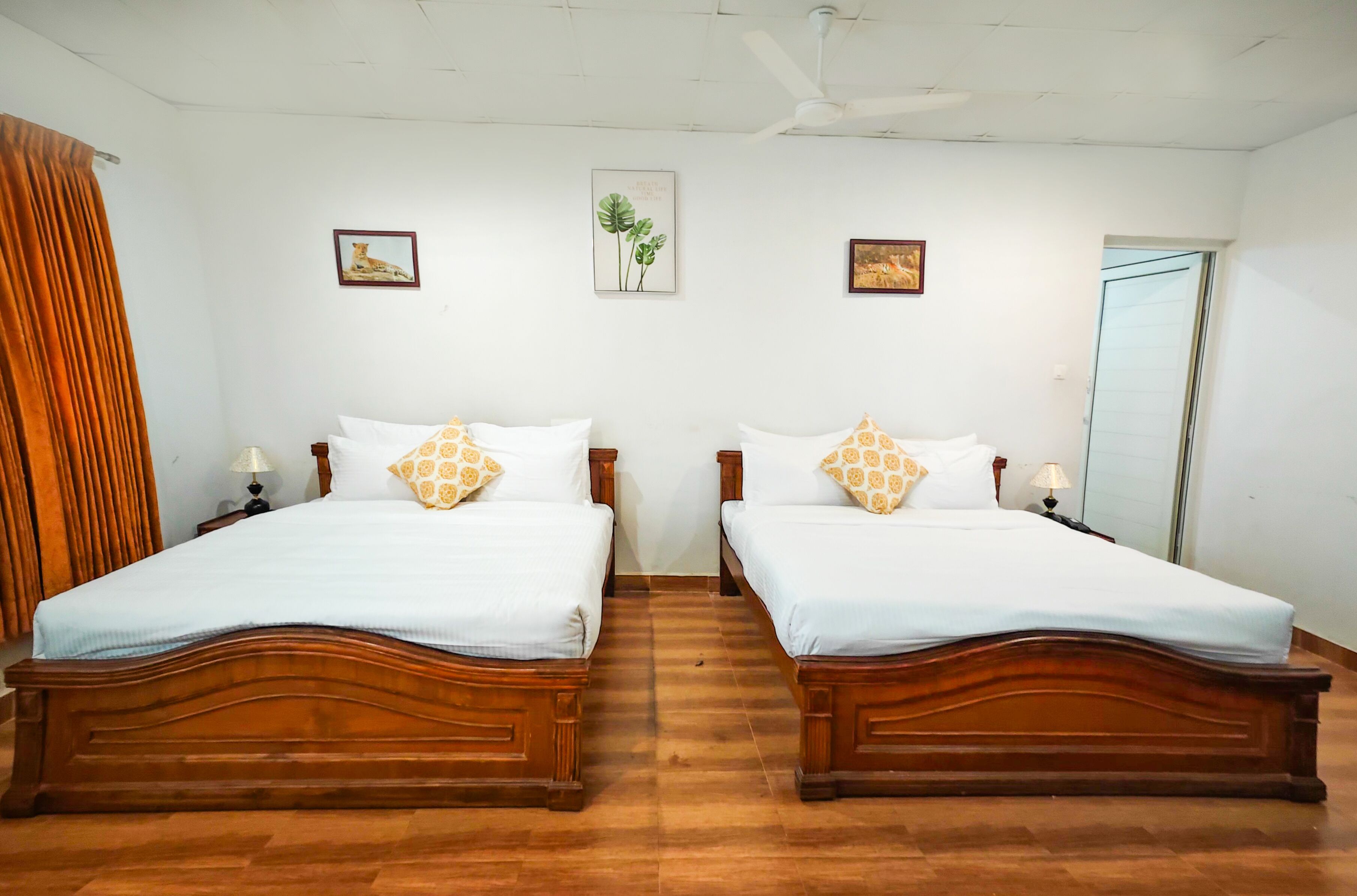 Una habitación con dos camas de madera, ropa de cama blanca y almohadas estampadas. Hay cuadros en la pared y un ventilador de techo.