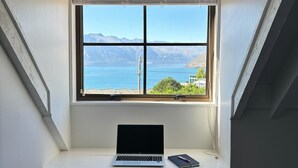 Ein Laptop auf einem Schreibtisch, mit Blick auf eine Berglandschaft und ein Gewässer durch ein Fenster.