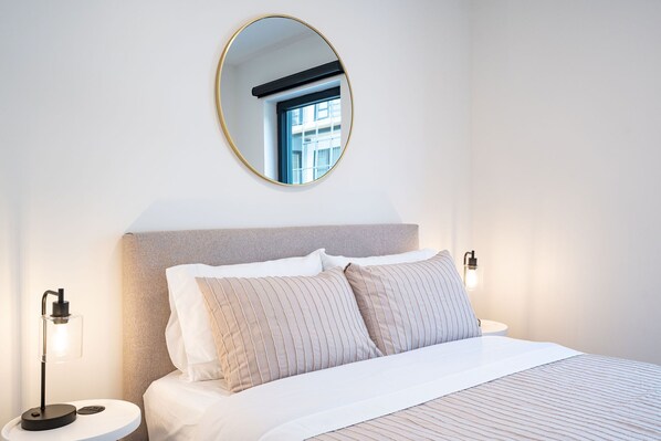 A neatly made bed with striped bedding, a round mirror on the wall, and a bedside lamp. - Mountainside condo with private hot tub & BBQ (Revelstoke)