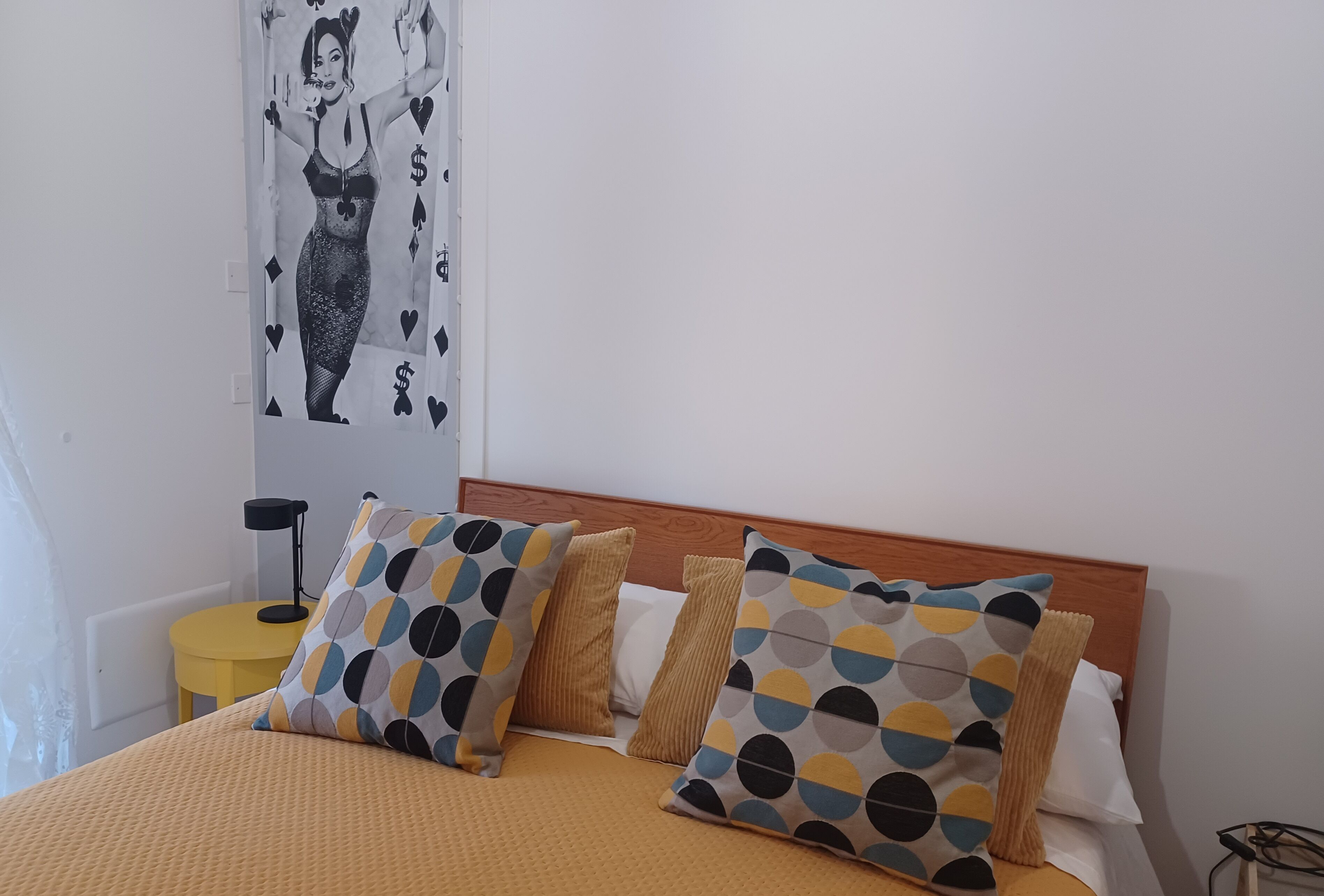 Ein Bett mit gelber Bettdecke, zwei gemusterte Kissen, ein hölzernes Kopfteil und ein kleiner gelber Tisch mit Lampe daneben.