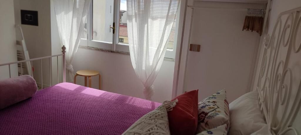 Quarto com cama, um banquinho pequeno e janela com cortinas leves.