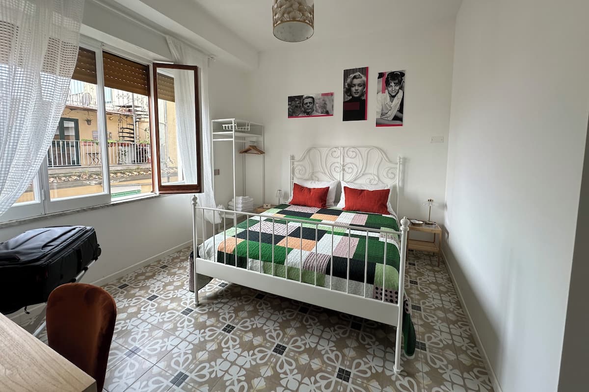 A bedroom with a white metal bed, a patterned floor, a window with curtains, and a hanging light fixture.