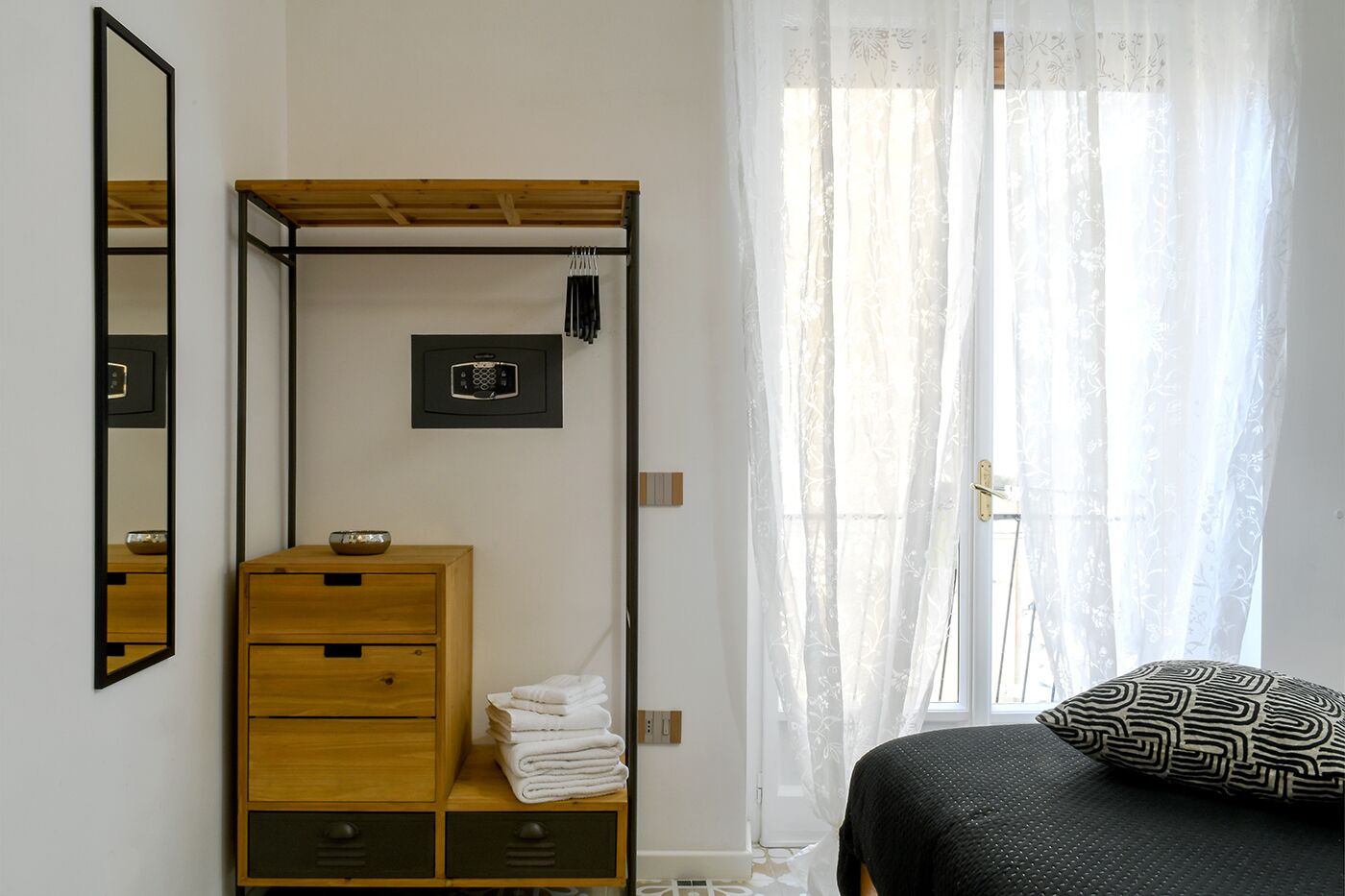 A bedroom with a wooden wardrobe, a bed with a patterned pillow, a mirror, and a window with sheer curtains.
