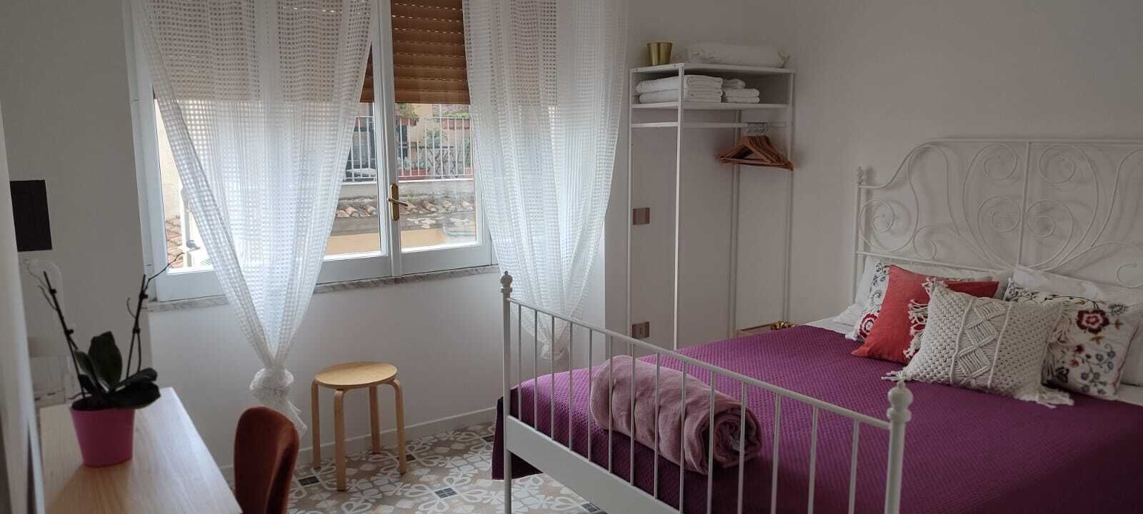 Ein Schlafzimmer mit einem Bett, einem Fenster mit Vorhängen, einem Nachttisch, einem Sessel und einem Kleiderschrank.