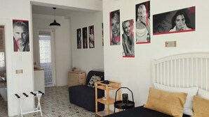 Un dormitorio con cama, mesita de noche y decoración en la pared. Se ve una puerta que da a otra habitación y una ventana con cortinas.