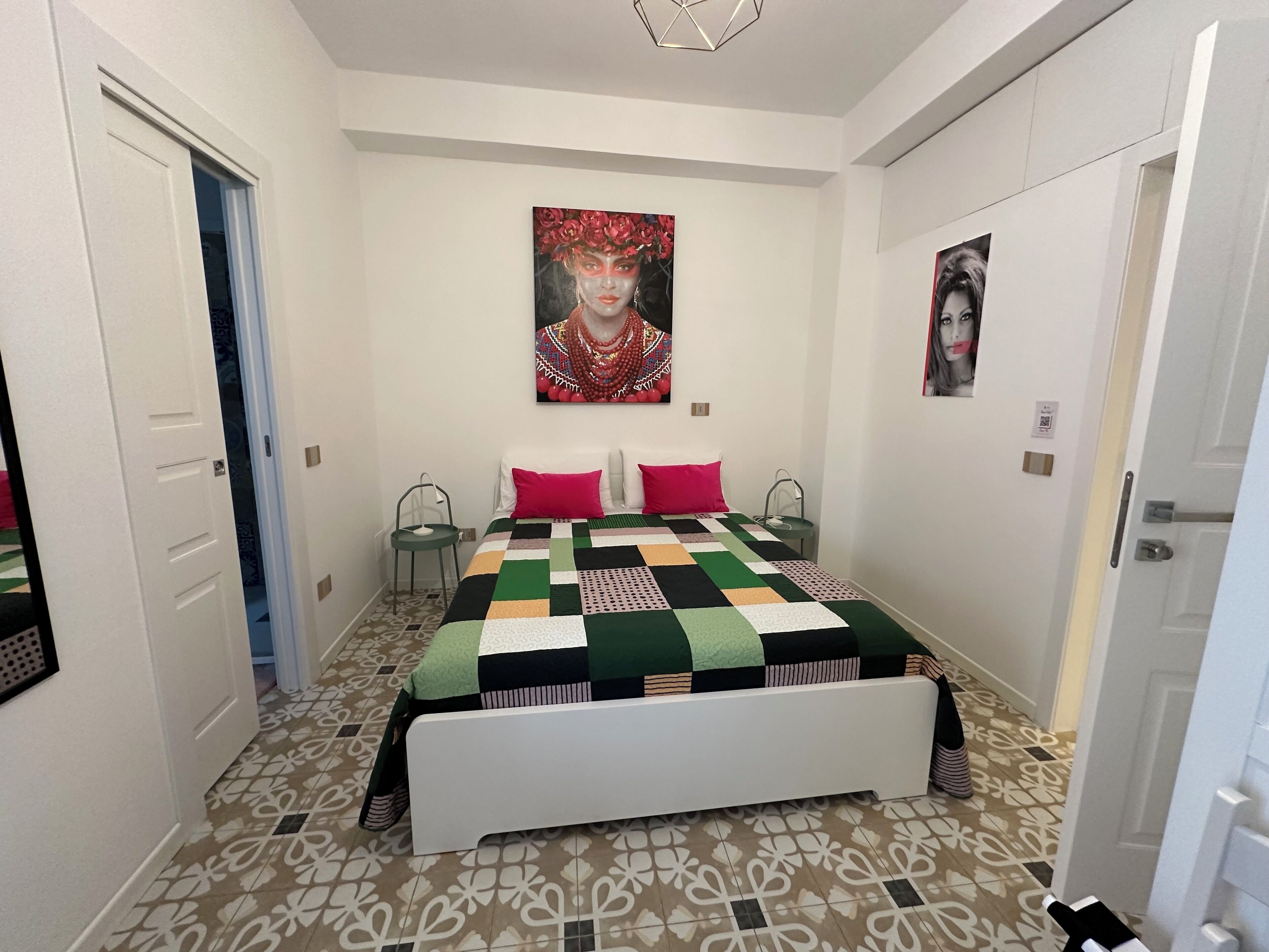 A bedroom with a bed, two chairs, a painting on the wall, and patterned floor tiles.