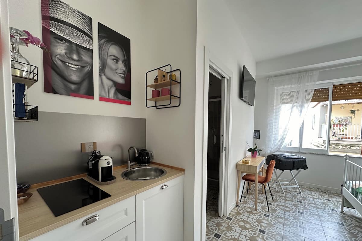 A modern kitchen with a sink, coffee maker, and a small table with a chair. There are decorative items on the wall and a window with a view outside.