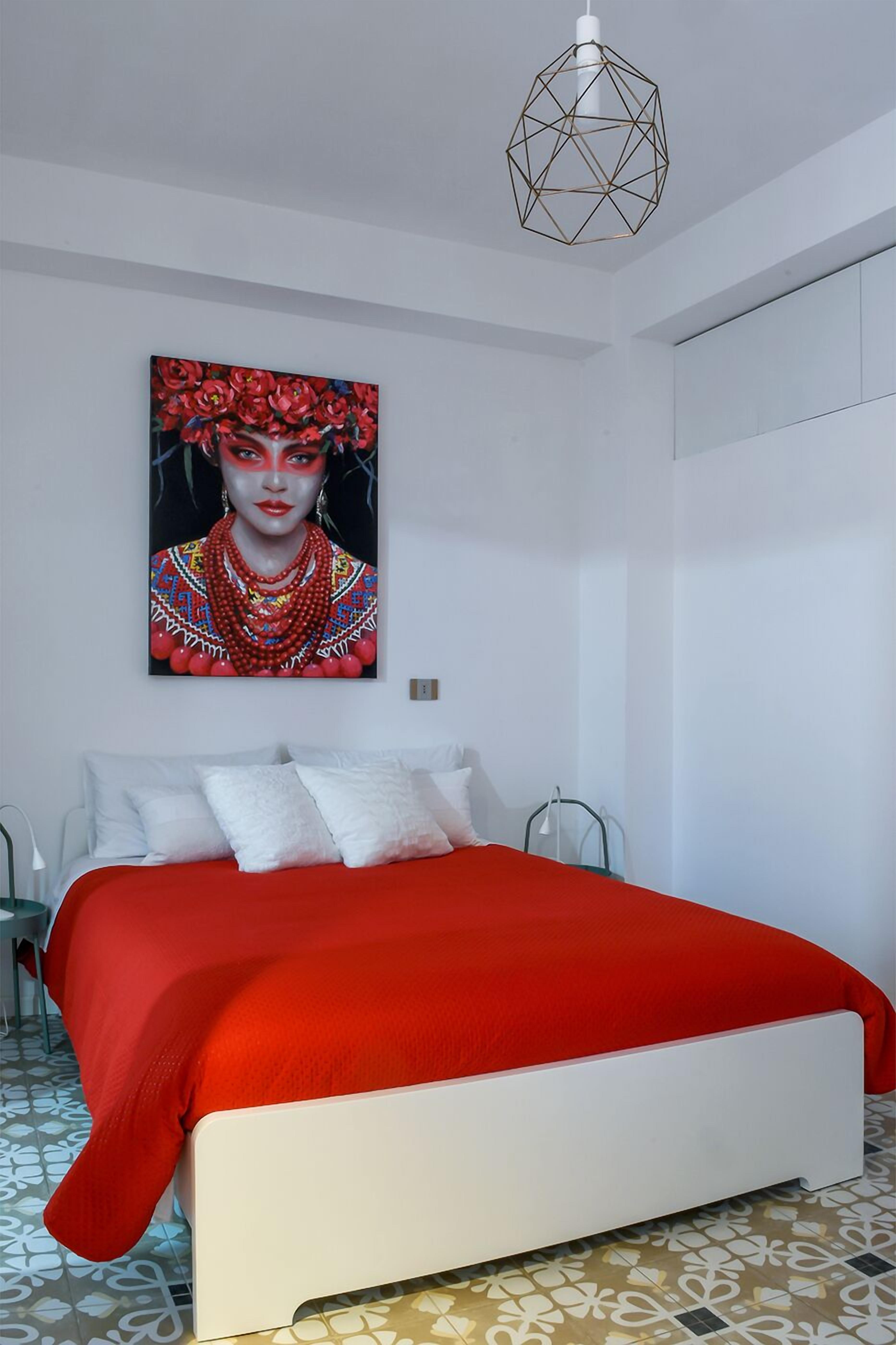 Un dormitorio moderno con cama blanca, ropa de cama roja, una lámpara colgante geométrica y un cuadro abstracto colorido en la pared.