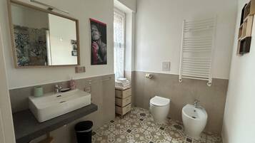 Un baño con un lavamanos blanco, un inodoro y un bidé. Se observa un espejo sobre el lavamanos, y un perchero para toallas. El piso tiene baldosas con un diseño.