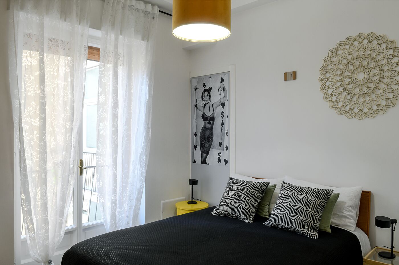 Une chambre à coucher avec un lit, des coussins à motifs, une œuvre d’art murale, une décoration murale et une fenêtre avec des rideaux légers.