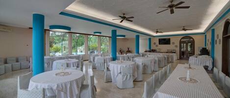 A spacious banquet hall with round tables and chairs, large windows, and ceiling fans.