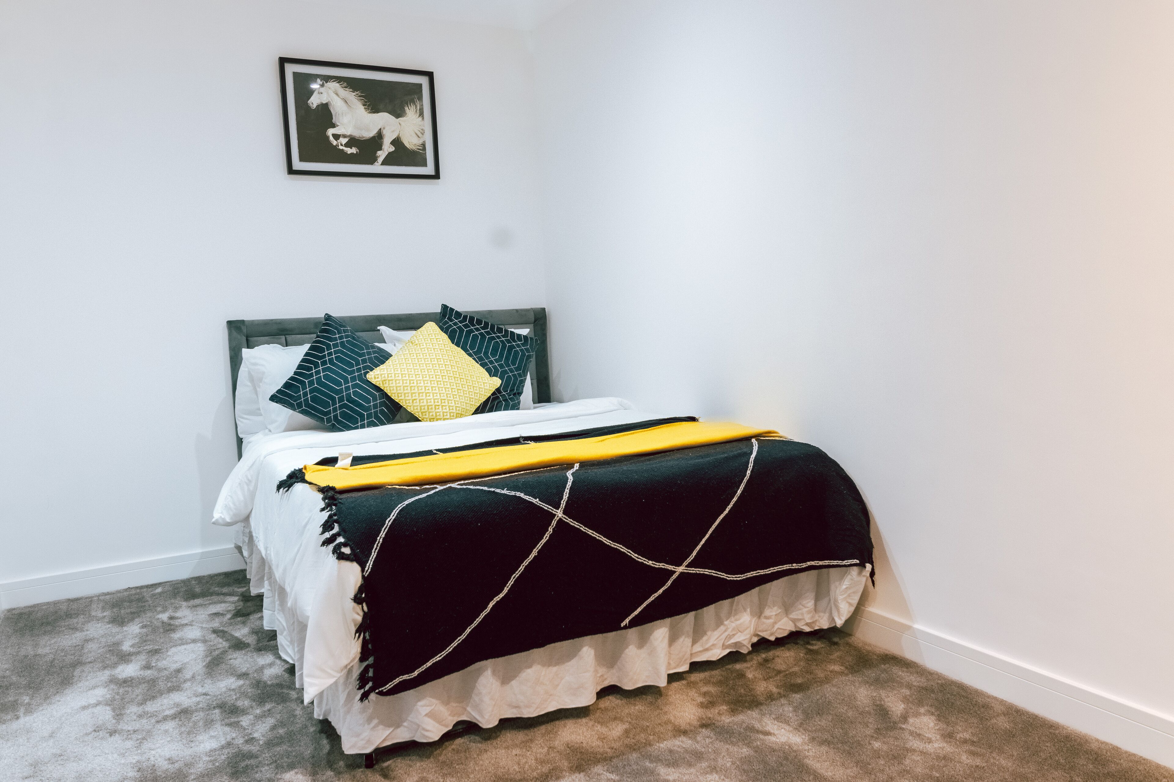 Une chambre à coucher avec un lit, des murs blancs, un tableau encadré et un sol recouvert de moquette.