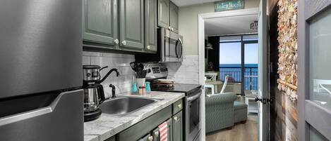 Une cuisine avec des meubles de couleur vert foncé, un réfrigérateur en acier inoxydable, une machine à café et un four à micro-ondes. On peut apercevoir un balcon avec une pancarte indiquant « Beach House ».