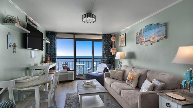 A living room with a view of the sea, a balcony, and a dining area with a TV. - Oceanfront Premier King Ste Free Breakfast 8th Flr (Myrtle Beach)