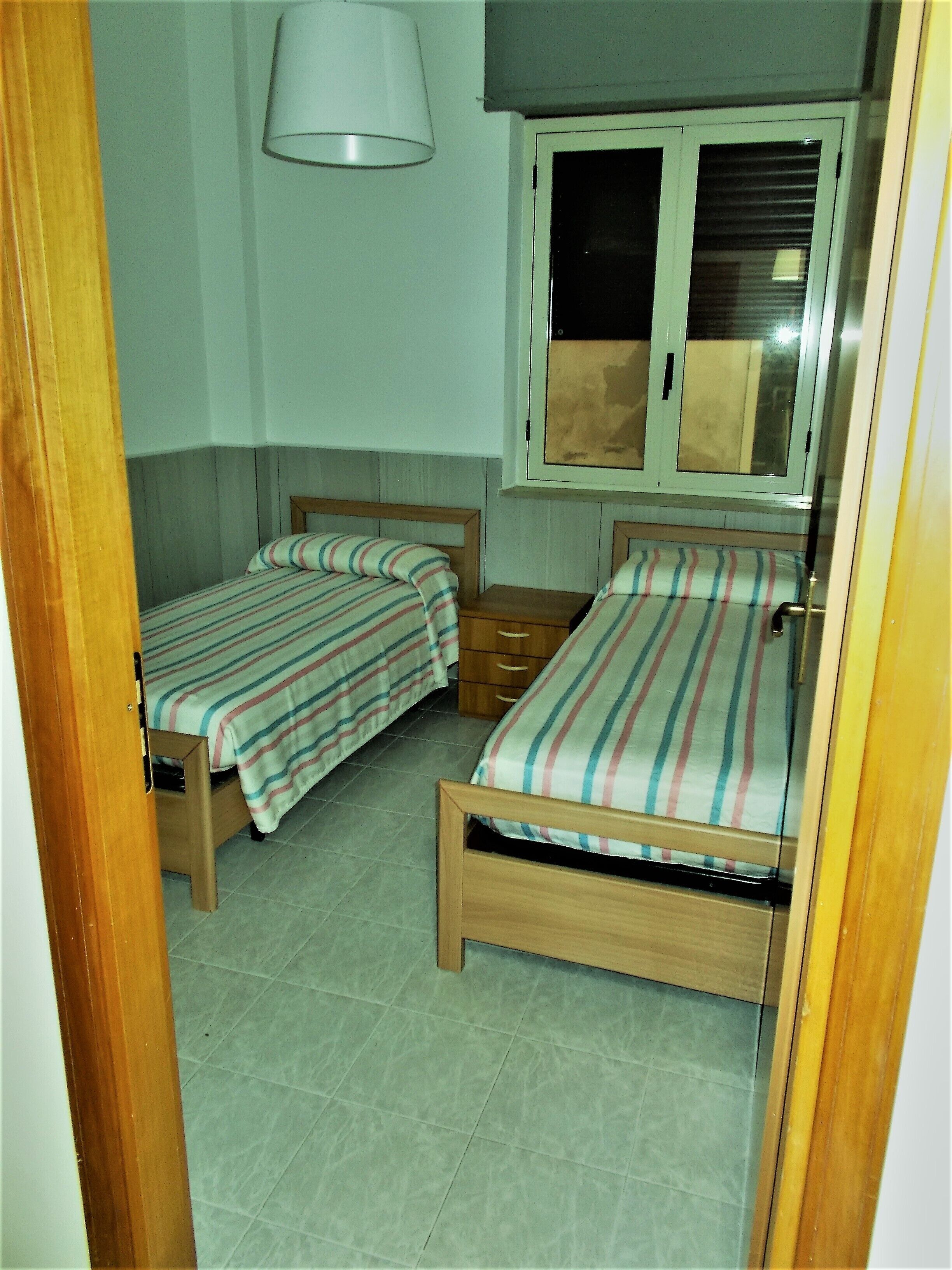 A bedroom with two single beds, a bedside table, a lamp, and a window with blinds.