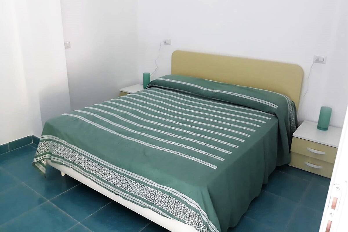 A single bed with a green striped bedspread and a patterned pillow, a wooden headboard, and a small bedside table with a green object on it.