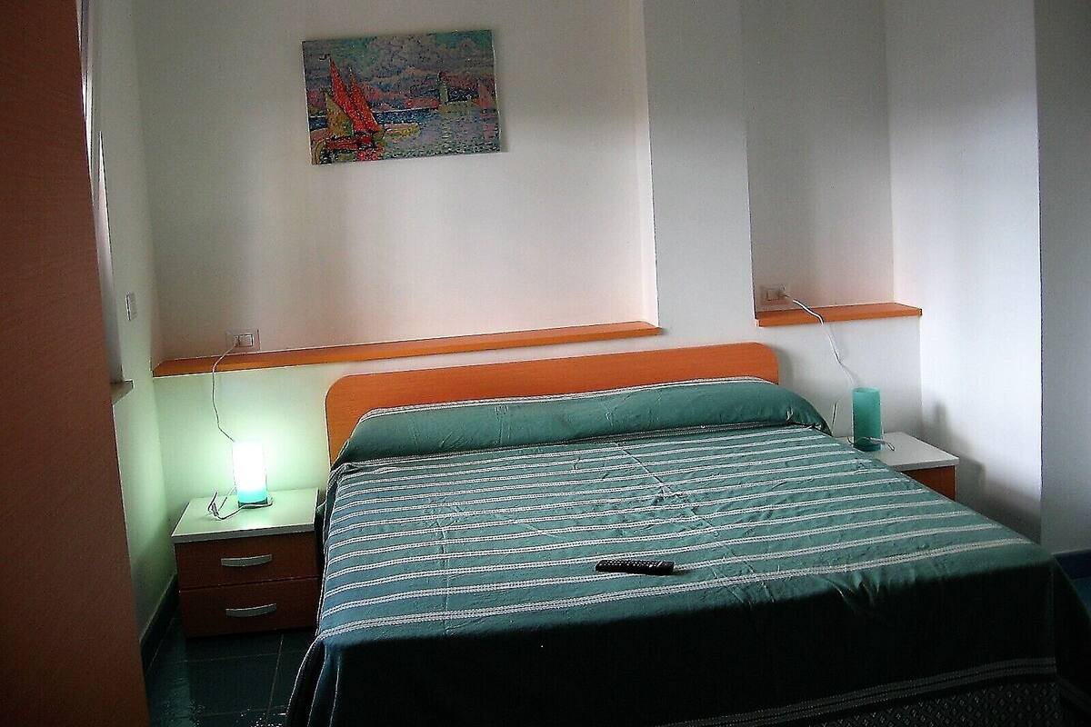 A bedroom with a bed, a nightstand, a wall-mounted shelf, and a painting.