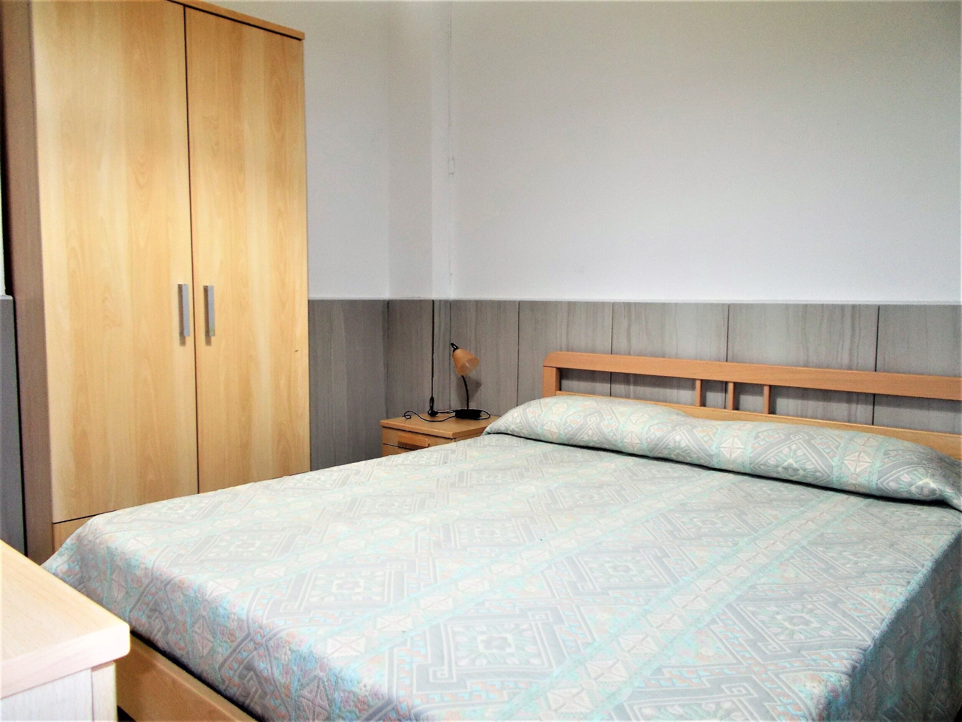 A bedroom with a bed, a wooden wardrobe, a nightstand, and a lamp.