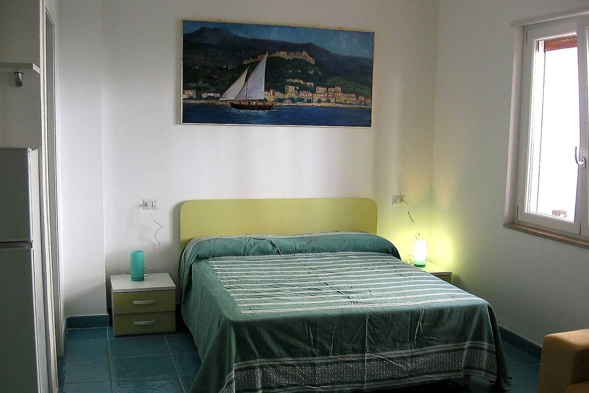 A bedroom with a bed, a nightstand, a lamp, and a painting of a coastal scene.