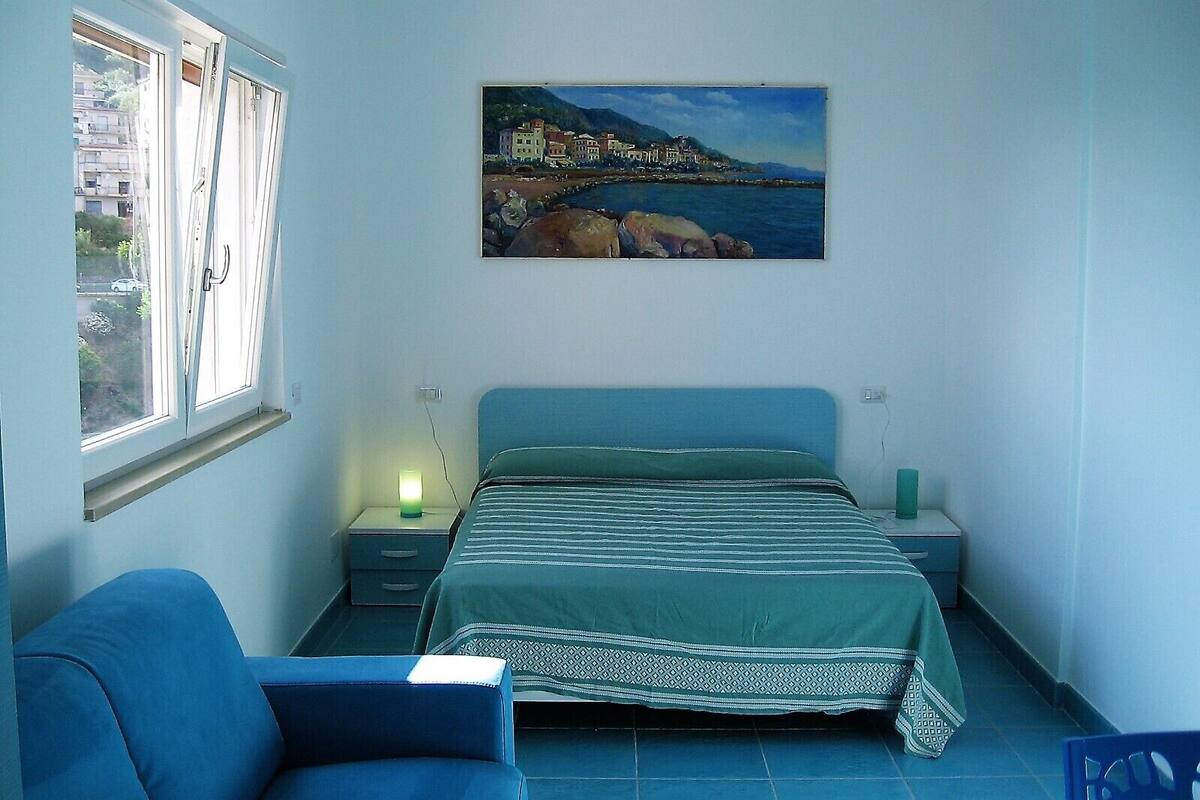 A bedroom with a bed, a chair, a nightstand with a lamp, and a painting of a coastal town on the wall.