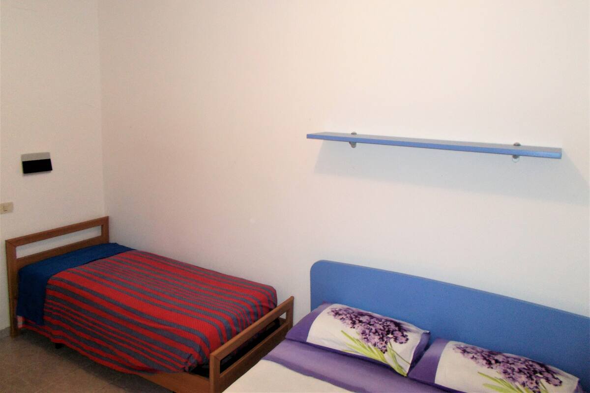 A small, simple room with two beds, one with a red and blue striped bedspread and the other with purple pillows and a white pillow with a purple floral design. There is a blue shelf on the wall above the beds.