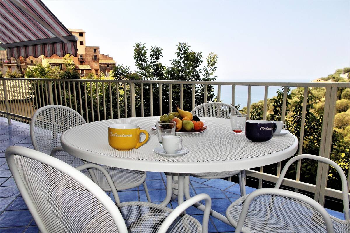 A round table with chairs on a balcony, a yellow mug, a bowl of fruit, and a Polazzio mug.