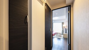 Un pasillo con una puerta de madera oscura, una pared de color claro y una vista parcial de una habitación con cama y lámpara.