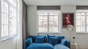 A room with a blue sectional sofa, a white coffee table, and a floor lamp. - Designer 1BR Soho Apt | AC | Heart of West End (London)