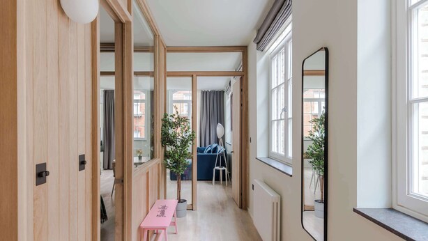 A corridor with wooden doors, a pink bench, a mirror, and a view of a blue sofa and curtains. - Designer 1BR Soho Apt | AC | Heart of West End (London)