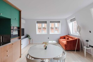 A modern apartment with a kitchen, dining area, and living room. There are two windows, a round dining table, and a sofa.