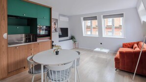 A modern kitchen with wooden cabinets, a dining area with a round table and chairs, a sofa, and two windows with a view of buildings. - Top-Floor Soho Flat | AC | Sleeps 4 | Central Ldn (Greater London)
