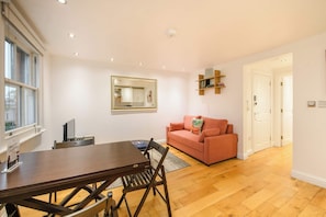 A spacious living room with a wooden floor, a dining table with chairs, a sofa, and a large mirror on the wall.