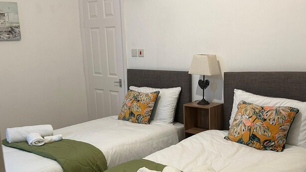 Ein Hotelzimmer mit zwei Betten, jeweils mit einer grünen Tagesdecke und weißen Bettwäsche. Es gibt einen hölzernen Nachttisch mit einer Lampe und ein dekoratives Kissen mit tropischem Muster.