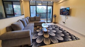 Un salon moderne avec un canapé d’angle, un tapis à motifs, une petite table, un téléviseur et une vue sur le balcon.