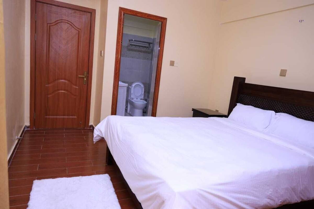A bedroom with a wooden door, a white bed, and a bathroom visible through a glass door.
