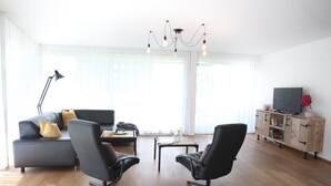 A modern living room with a sofa, two black chairs, a coffee table, a TV on a wooden cabinet, and a chandelier.